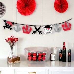 May include: A pirate-themed party banner with red and white ships, skulls, and crossbones. The banner is strung on a black string and is hanging on a white wall. There are two red paper fans on the wall behind the banner.