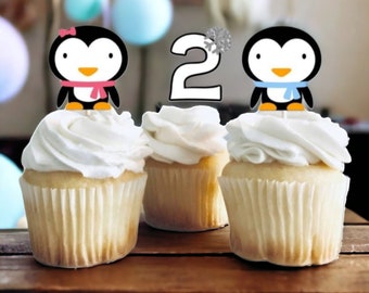 May include: Three cupcakes decorated with penguin and number two toppers. The penguins are wearing scarves and hats. The cupcakes are frosted with white frosting.