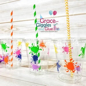 May include: Four clear plastic cups with colourful paint splatter designs. Each cup has a straw, one orange and white, one green and white, and two yellow and white. The cups are on a wooden surface. The text on the cups reads "Grace Giggles and Glue" and "Custom party and event decor".
