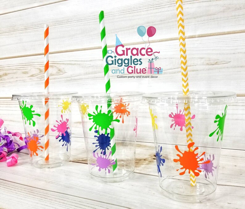 May include: Four clear plastic cups with colourful paint splatter designs. Each cup has a straw, one orange and white, one green and white, and two yellow and white. The cups are on a wooden surface. The text on the cups reads "Grace Giggles and Glue" and "Custom party and event decor".