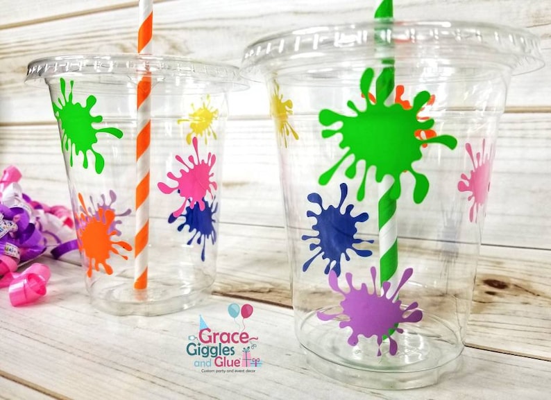 May include: Two clear plastic cups with colourful paint splatter designs. The cups have orange and white striped straws. The cups are on a white wooden background.