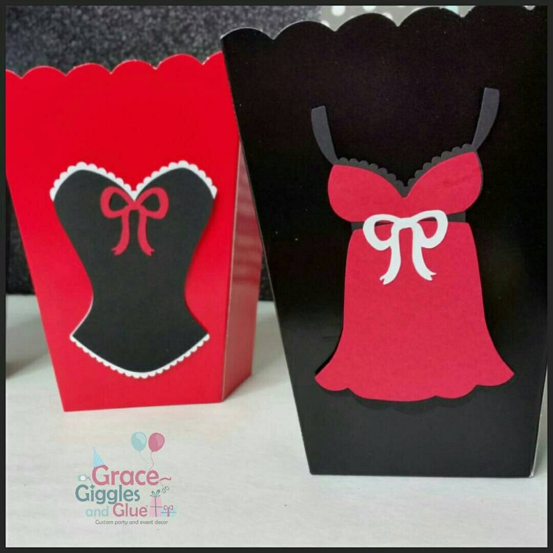 May include: Two party favor boxes, one red and one black, with a black and white corset and a red dress cutouts. The boxes have a scalloped edge and are decorated with a white bow.
