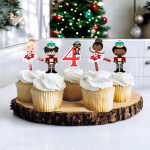 May include: Six cupcakes decorated with a Nutcracker theme. The cupcakes are topped with a red and white number four, a ballerina with a red dress, a Nutcracker with a blue uniform, and a Nutcracker with a red uniform.
