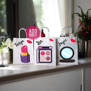 May include: Three white paper gift bags with handles. Each bag has a different makeup-themed design: a lipstick, an eyeshadow palette, and a compact mirror. The bags are labeled with the names "Tanya", "Lia", and "Joyce".