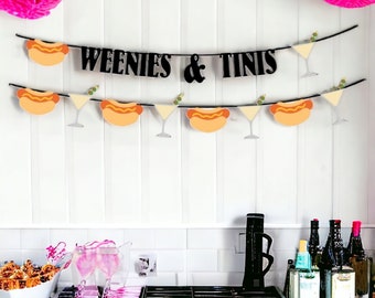 May include: A party banner featuring black text that reads WEENIES & TINIS, accompanied by alternating cutouts of hot dogs and martini glasses with olives. The banner is strung on a black cord and accented with bright pink paper pom-poms.