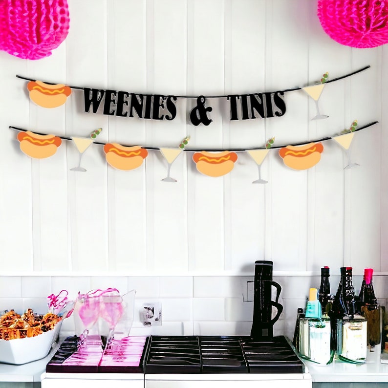 May include: A party banner featuring black text that reads WEENIES & TINIS, accompanied by alternating cutouts of hot dogs and martini glasses with olives. The banner is strung on a black cord and accented with bright pink paper pom-poms.