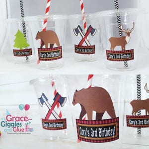 May include: Clear plastic cups with colorful paper designs featuring a bear, a deer, a tree, and crossed axes. The cups have red and white striped straws and a red and black plaid banner with the text "Cory's 3rd Birthday!"