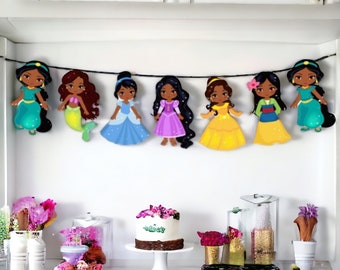 May include: A colorful banner with seven princess characters, each wearing a different dress and hairstyle. The banner is strung on a black cord and is hanging from a white cabinet. There is a cake with pink flowers on top on a white cake stand in the foreground.
