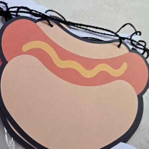 May include: A decorative paper hot dog cutout featuring a tan bun, a reddish-brown sausage, and a wavy yellow mustard line. The cutout has a black border and is strung together with black yarn, forming part of a hanging garland banner.