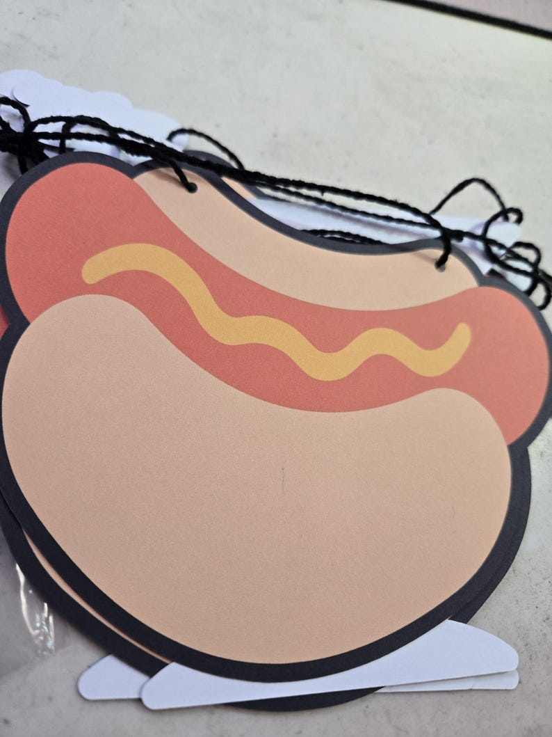 May include: A decorative paper hot dog cutout featuring a tan bun, a reddish-brown sausage, and a wavy yellow mustard line. The cutout has a black border and is strung together with black yarn, forming part of a hanging garland banner.