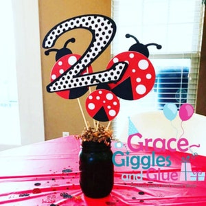 May include: A black jar centerpiece with ladybug and number two cutouts. The number two is white with black polka dots. The ladybugs are red with white spots and black accents. The jar sits on a red tablecloth. The text "Grace Giggles and Glue" is visible.