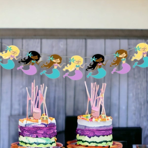 May include: A mermaid-themed party banner with seven colorful mermaids, each with a different hair color and tail. The banner is strung between two cakes decorated with purple frosting and colorful sprinkles.