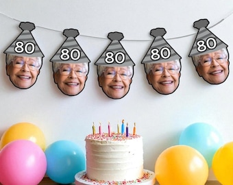 May include: A festive birthday scene with a white cake adorned with colorful sprinkles and lit candles. Above, a banner of smiling faces wearing party hats with the number 80. Colorful balloons in pink, yellow, and blue complete the celebratory setting.