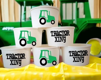 May include: A stack of white paper cups with green tractor designs and the text "Tractor Xing" on them. The cups are stacked on a yellow tablecloth.