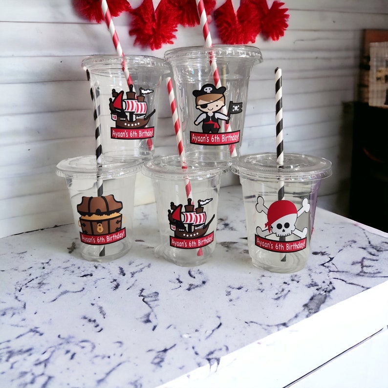 May include: Six clear plastic cups with red and white striped straws. Each cup has a personalized sticker with a pirate theme and the text "Ayaan's 6th Birthday".