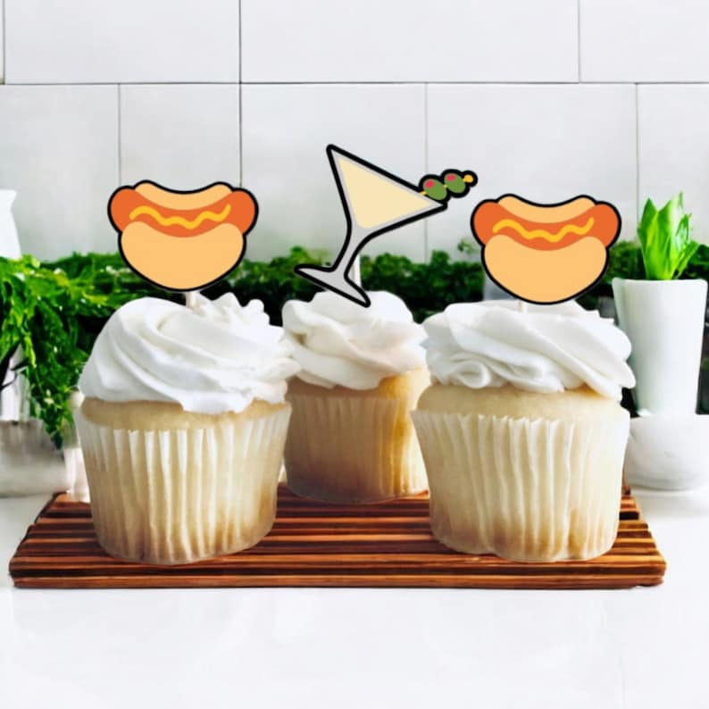 May include: Three cupcakes with white frosting, each topped with a decorative paper topper. Two toppers feature a hot dog illustration, and the center topper displays a martini glass with two olives on a toothpick.