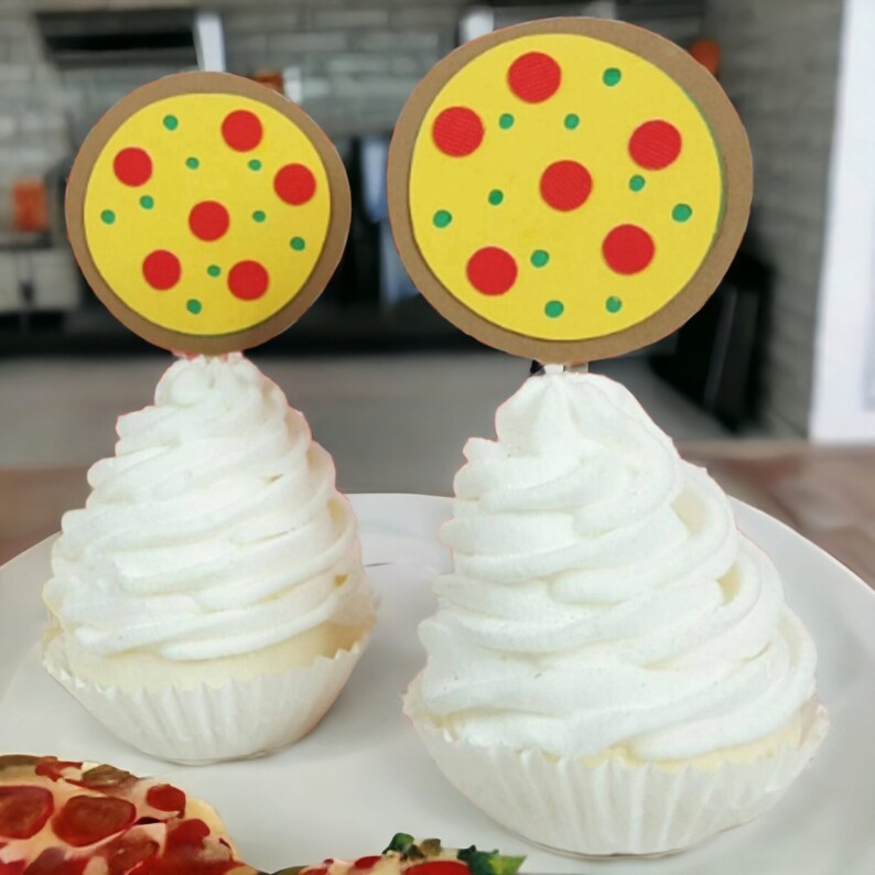 Puede incluir: Dos cupcakes blancos con un remolino de glaseado cubiertos con una rebanada de pizza de papel con ingredientes rojos y verdes.
