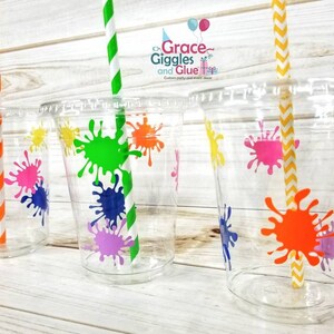 May include: Four clear plastic cups with colourful paint splatter designs. Each cup has a striped straw inserted. The cups are on a light wood surface.