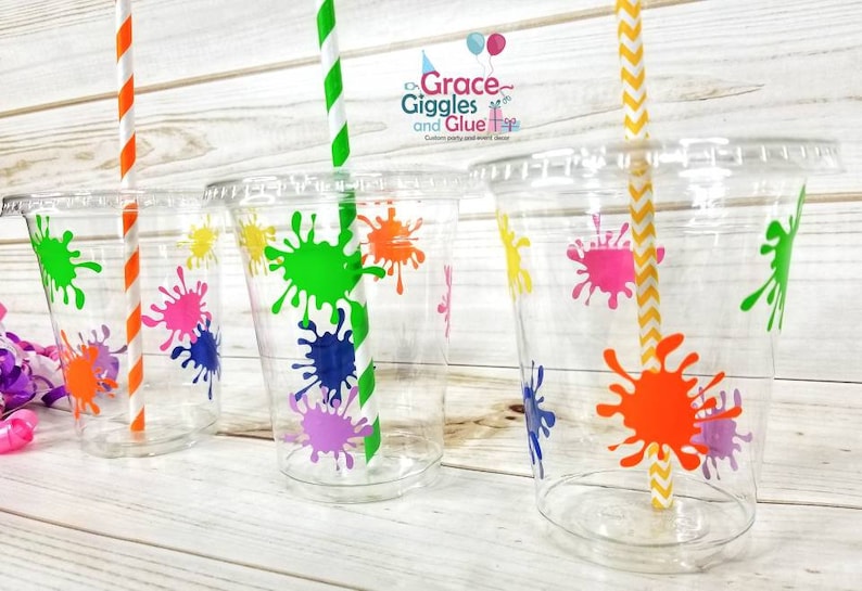 May include: Four clear plastic cups with colourful paint splatter designs. Each cup has a striped straw inserted. The cups are on a light wood surface.