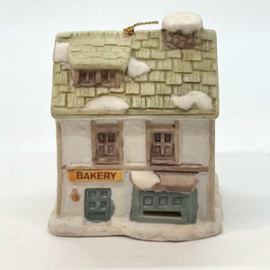 May include: A ceramic miniature bakery ornament with a tan roof, white walls, and a sign that reads "BAKERY." The ornament has a gold string for hanging and is decorated with snow accents. The windows and door are painted in shades of brown and blue.
