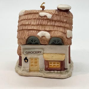 May include: A ceramic miniature grocery store figurine with a thatched roof, snow accents, and the word "GROCERY" above the entrance. The building has a yellow door and window, and a small light fixture. The figurine is on a white surface.