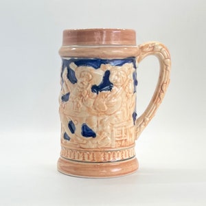 May include: A ceramic beer stein with a brown and blue glaze. The stein features a raised design of people playing music and drinking.