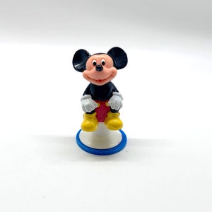 May include: A black and red Mickey Mouse figurine sitting on a white and blue base. The figurine is made of plastic and has a cartoonish style.