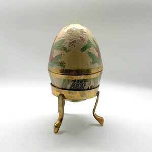 May include: A decorative egg-shaped trinket box with a gold stand. The egg is painted with floral designs in shades of green, red, and white.