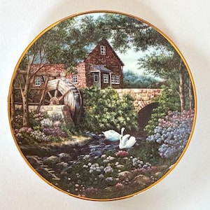 May include: Decorative plate featuring a scenic landscape with a water mill, bridge, and two swans in a stream. The plate has a gold rim and a detailed painting of a rural scene with lush greenery and a brick building.