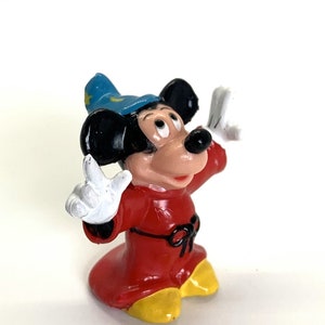 May include: A small plastic figurine of Mickey Mouse dressed as a sorcerer. He is wearing a red robe with a black belt, a blue pointed hat with yellow stars, and white gloves. He has a surprised expression on his face and his arms are raised.