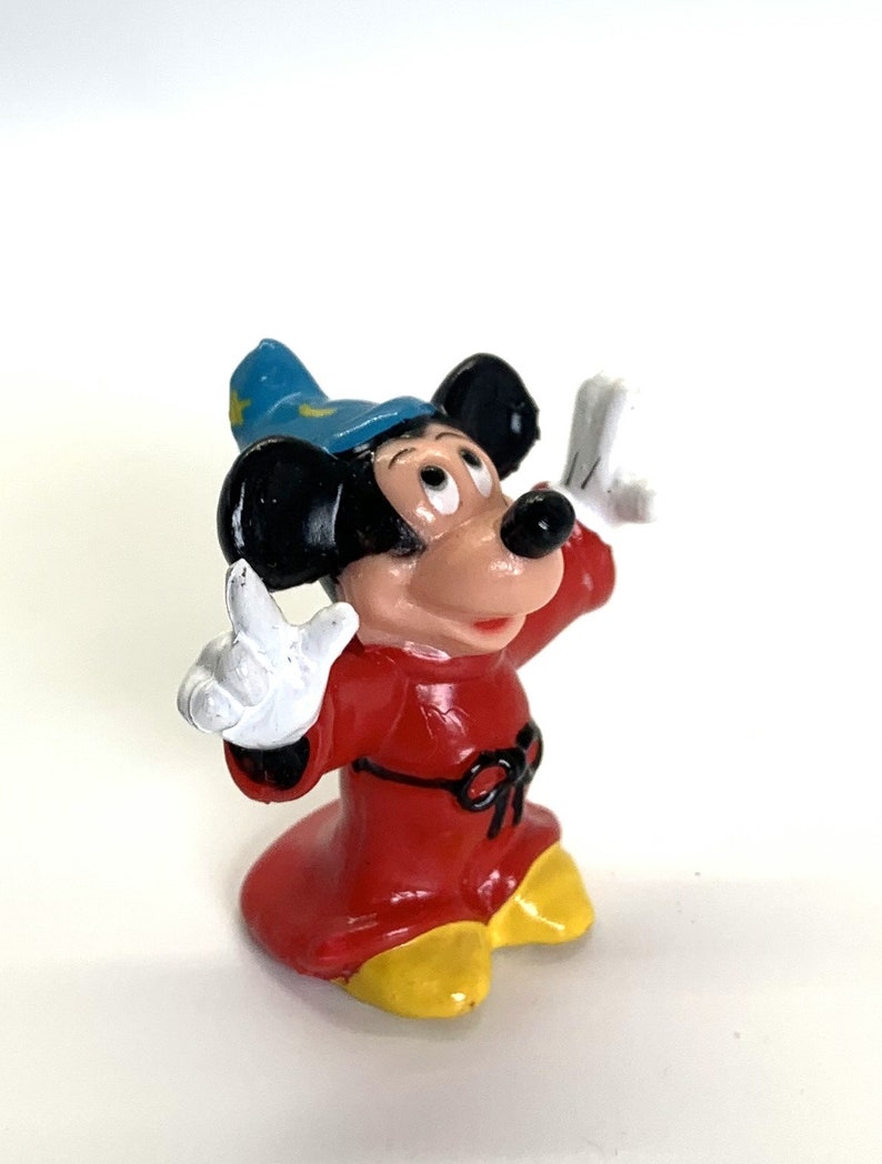 May include: A small plastic figurine of Mickey Mouse dressed as a sorcerer. He is wearing a red robe with a black belt, a blue pointed hat with yellow stars, and white gloves. He has a surprised expression on his face and his arms are raised.