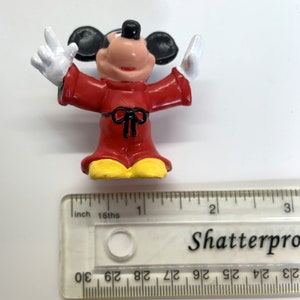 May include: A red and yellow plastic figurine of Mickey Mouse wearing a red robe with a black bow. The figurine is about 2 inches tall.
