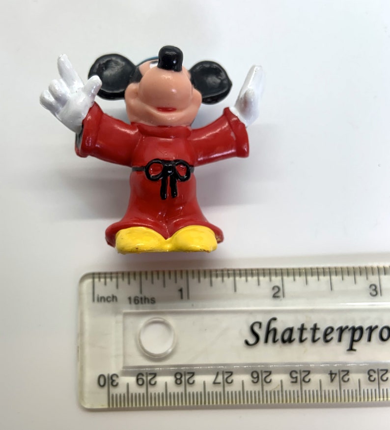May include: A red and yellow plastic figurine of Mickey Mouse wearing a red robe with a black bow. The figurine is about 2 inches tall.