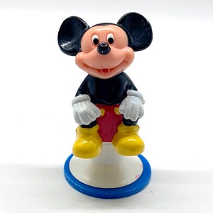 May include: A small plastic figurine of Mickey Mouse sitting on a white base with a blue ring. Mickey is wearing his signature red shorts, yellow shoes, and white gloves. He has black ears and a black body.