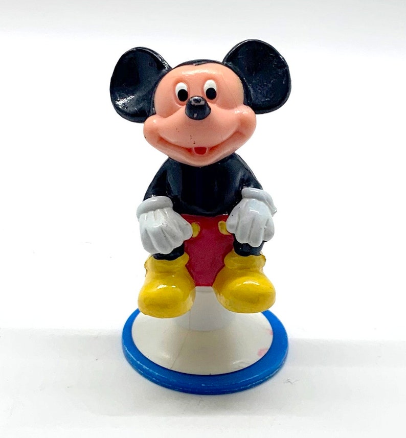 May include: A small plastic figurine of Mickey Mouse sitting on a white base with a blue ring. Mickey is wearing his signature red shorts, yellow shoes, and white gloves. He has black ears and a black body.