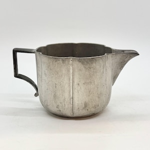 May include: A vintage silver-colored creamer pitcher with a scalloped rim and a spout. The pitcher has a dark handle and vertical lines on the body. The creamer is likely made of metal.