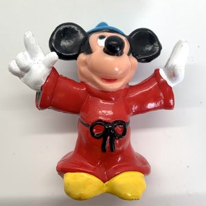 May include: A red and black sorcerer Mickey Mouse figurine with a blue hat. Mickey is wearing a red robe with a black bow and has yellow shoes.
