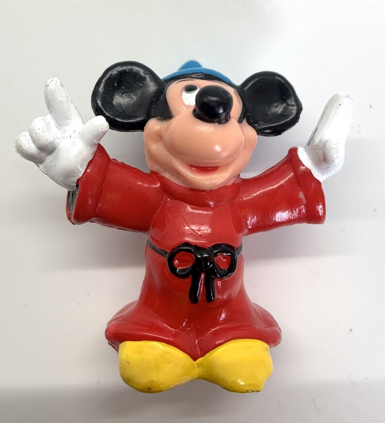 May include: A red and black sorcerer Mickey Mouse figurine with a blue hat. Mickey is wearing a red robe with a black bow and has yellow shoes.