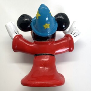 May include: A red and blue plastic figurine of Mickey Mouse dressed as a wizard. He is wearing a blue hat with yellow stars and a red robe. He is holding his arms out in a gesture of magic.
