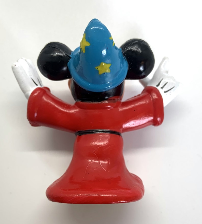 May include: A red and blue plastic figurine of Mickey Mouse dressed as a wizard. He is wearing a blue hat with yellow stars and a red robe. He is holding his arms out in a gesture of magic.