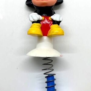May include: A spring-loaded toy featuring a cartoon Mickey Mouse figure. Mickey is sitting with his hands on his lap, wearing red shorts and yellow shoes. The toy has a white base and a blue spring.