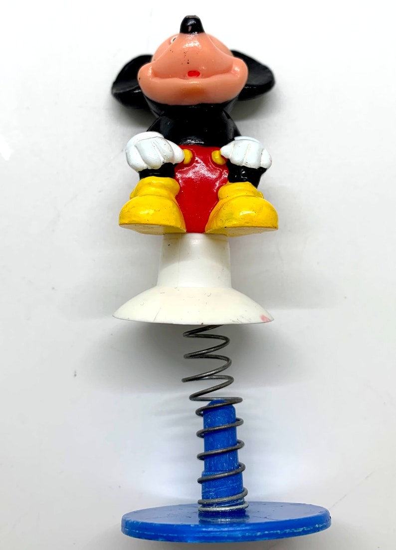 May include: A spring-loaded toy featuring a cartoon Mickey Mouse figure. Mickey is sitting with his hands on his lap, wearing red shorts and yellow shoes. The toy has a white base and a blue spring.