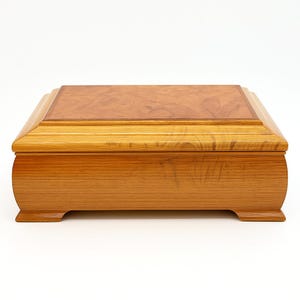 May include: A rectangular wooden box with a light brown finish. The lid features a darker, patterned wood inlay. The box has rounded sides and sits on small feet. The overall design is classic and elegant, suitable for storage.