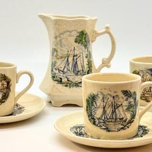 May include: A cream-colored porcelain tea set featuring a pitcher and three cups with saucers. Each piece is decorated with a nautical scene in blue, green, and brown. The set is displayed on a white background.