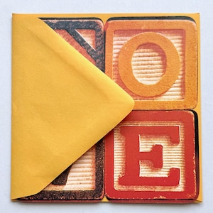 May include: A greeting card with the word "NOTE" spelled out in colorful, vintage-style alphabet blocks. The blocks are orange and red with a distressed look, and a yellow envelope partially covers the card.