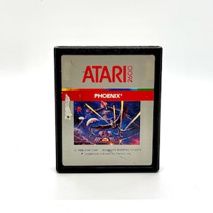 May include: A black Atari 2600 game cartridge with a silver label. The label features the text "ATARI 2600" in white and "PHOENIX*" in red. The game cartridge has a colorful image of a spaceship in space.