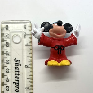 May include: A red and yellow plastic figurine of Mickey Mouse dressed as a sorcerer. He is wearing a red robe with a black sash and a pointed hat. His arms are outstretched and his hands are in a gesture of casting a spell.