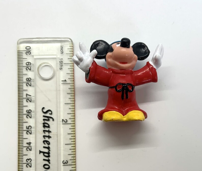 May include: A red and yellow plastic figurine of Mickey Mouse dressed as a sorcerer. He is wearing a red robe with a black sash and a pointed hat. His arms are outstretched and his hands are in a gesture of casting a spell.