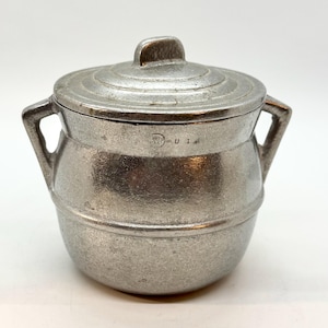 May include: A small, silver-colored metal pot with a lid and two handles. The pot has a rounded body and a lid with a small handle on top. The pot is marked with the letters "U S A".