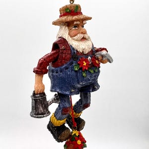 May include: A hanging ornament of a bearded figure wearing a straw hat, red plaid shirt, and blue denim overalls with red poinsettia accents. The figure holds a silver watering can and is connected by a red string to a small potted poinsettia.